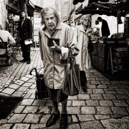 Two elderly women in trench coats walk through a cobblestone marché. One stands in the foreground holding a handbag and wallet, her expression concerned. Market stalls and people bustle in the background.