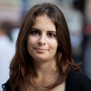 A woman with long brown hair is looking towards the camera with a neutral expression. She has a light complexion and is wearing a gold necklace with a small pendant and a dark top. The blurred background hints at an outdoor scene, possibly capturing the essence of the 2009 Techno Parade.