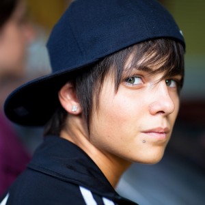 A person with short dark hair, wearing a black baseball cap backwards, looks over their shoulder at the 2009 Techno Parade. They have a subtle piercing on the lower lip and are dressed in a dark jacket with white stripes on the shoulders. The background is out of focus, adding to the vibrant atmosphere.