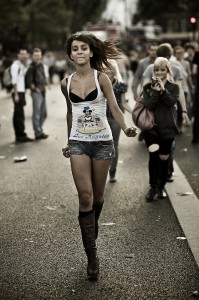 A woman with long hair walks confidently down a street, wearing a white tank top with a design, denim shorts, and knee-high boots. She is amidst a lively parade of people, some walking and others standing in the background. The atmosphere buzzes like a 2009 techno festival.