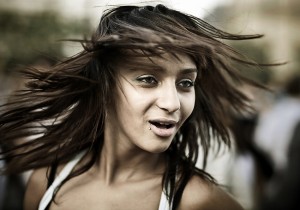 A person with long brown hair is in motion, their hair flying around their face. They have a small piercing below their lower lip and appear to be outdoors at the 2009 Techno Parade. The background is blurred, suggesting movement and capturing their expressive facial features.