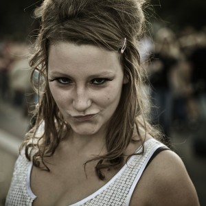 A woman with wavy brown hair and a hair clip poses with a playful, pouty expression. She is wearing a white top with a circular pattern. The background appears to be an outdoor event, perhaps the 2009 Techno Parade, with blurred people and greenery.