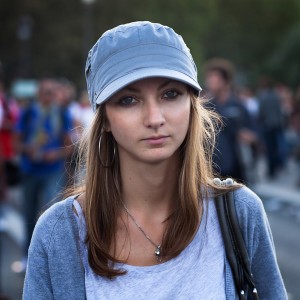 A young woman with long brown hair is wearing a grey cap, hoop earrings, and a casual grey outfit, reminiscent of 2009 fashion. She is outdoors at the Techno Parade with other people blurred in the background. Her expression is neutral, and she appears to be looking directly at the camera.