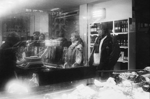 Black and white photo of people waiting inside a grocery store in Ostende. They stand in front of a counter filled with food items, and shelves with bottles behind them. The reflection on the glass adds a layer of texture to the scene, capturing a quiet errance in their wait.