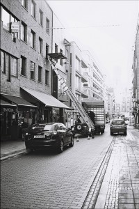 Black and white photo of a city street in Ostende, where a ladder truck is positioned next to a building. Workers appear to be accessing an upper floor through a window. Cars are parked along the street, and shops with signs are visible, including a tea room named Errance.