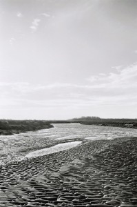 A black and white photograph of a riverbed during low tide, showcasing rippled patterns in the wet sand. The river meanders toward the horizon, capturing a sense of errance, where distant trees are visible beneath a partly cloudy sky.