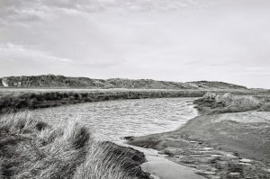 A black and white photograph capturing a tranquil landscape with a winding river flowing through a grassy area in Ostende. Low hills are visible in the distance under a partly cloudy sky. The water and grasses create a peaceful, natural scene, evoking an air of errance through untouched beauty.