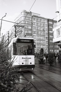 A black and white photograph of a tram labeled "Kusttram" heading to its destination "Knokke." The scene, possibly captured during an errance from Ostende, features a few pine trees and multi-story city buildings. The wet street suggests recent rain, with people visible in the distance.