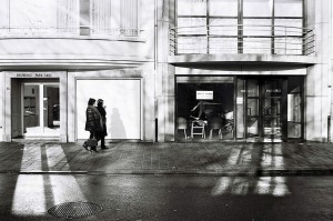 A black-and-white photo of two people walking on a sidewalk lined with buildings in Ostende. They pass by closed and open shops with large windows. Shadows of tree branches are cast on the buildings and the wet sidewalk, capturing a sense of errance.