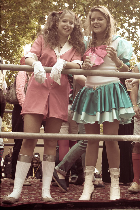 Two women in costumes stand behind a metal railing at the 2010 Techno Parade. One wears a pink outfit with white gloves and knee-high socks, while the other dons a teal and white dress with white boots. Both are smiling amid the crowded outdoor setting, with trees visible in the background.