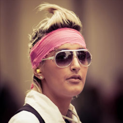 A person with blonde hair styled upwards, wearing a pink headband, white-framed sunglasses, and a lip piercing is seen in the image. They have earplugs in both ears and are dressed in a white shirt. The background is blurred, focusing attention on the person at the 2010 Techno Parade.