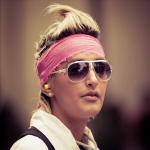A person with blonde hair styled upwards, wearing a pink headband, white-framed sunglasses, and a lip piercing is seen in the image. They have earplugs in both ears and are dressed in a white shirt. The background is blurred, focusing attention on the person at the 2010 Techno Parade.