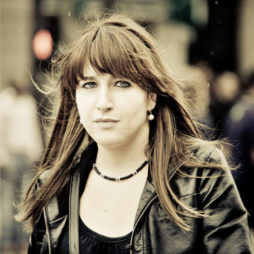 A young woman with long brown hair and bangs stands outdoors at a 2010 Techno Parade, wearing a black leather jacket, a black top, and a beaded necklace. She has a slight smile and appears to be looking directly at the camera. The background is blurred with a few indistinct people.