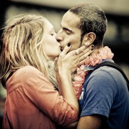 A man and woman share a passionate kiss during the 2010 Techno Parade. The man is wearing a blue shirt and a pink lei around his neck, while the woman has blonde hair and wears an orange top. They are standing close, her hand gently touching his face, against a blurred background full of vibrant energy.