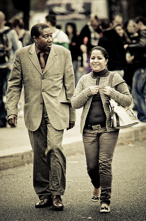 A man and a woman are walking outdoors in 2010. The man wears a gray suit and tie, while the woman is dressed in a cardigan, jeans, and holds a handbag. They are engaged in conversation with people and the blurred background of what appears to be a lively techno parade visible.