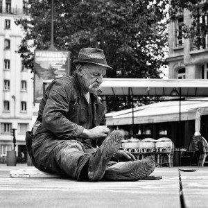 An older man in work clothes and a hat, perhaps a maître de la craie, sits on the ground, painting or working on a surface in an urban outdoor setting with buildings and a café in the background.