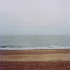 A wide, empty sandy beach stretches to the calm mer en hiver under an overcast sky; small gentle waves reach the shore, and the horizon divides the muted tones of water and sky.