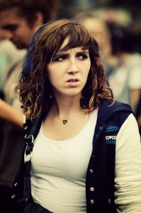 A young woman with curly brown hair, wearing a white top and a dark blue jacket, looks to the side with a concerned expression. She is in an outdoor, crowded setting at the 2011 Techno Parade music festival.