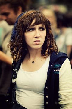 A young woman with curly brown hair, wearing a white top and a dark blue jacket, looks to the side with a concerned expression. She is in an outdoor, crowded setting at the 2011 Techno Parade music festival.