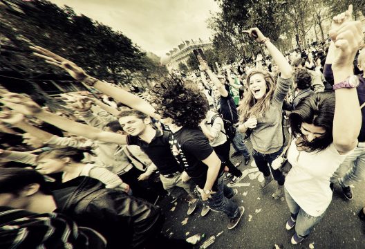 A vibrant crowd of people, mostly young, is energetically dancing and cheering at the 2011 Techno Parade. Arms are raised, and some individuals have wide smiles, capturing a moment of collective joy and excitement. Trees and a cloudy sky are visible in the background.