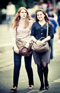 Two women walking side by side on a street, both carrying brown handbags. The one on the left with red hair is wearing a striped long-sleeve shirt and jeans, reminiscent of 2011's fashion. The one on the right with brown hair is dressed in a black cardigan and a gray skirt with tights, as if heading to a Techno Parade.
