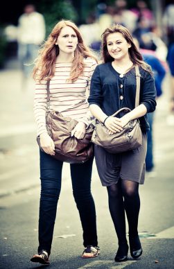 Two women walking side by side on a street, both carrying brown handbags. The one on the left with red hair is wearing a striped long-sleeve shirt and jeans, reminiscent of 2011's fashion. The one on the right with brown hair is dressed in a black cardigan and a gray skirt with tights, as if heading to a Techno Parade.