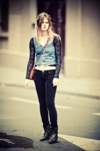 A woman with long hair stands on a city street looking forward. She is wearing a patterned black long-sleeve top underneath a blue sleeveless vest, paired with black pants and black boots. A brown crossbody bag is slung over her shoulder. The blurred background hints at the energy of the 2011 Techno Parade.