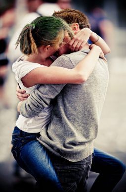 A person with green hair and a white shirt embraces another in a grey sweater, legs wrapped around their waist, seemingly taken during the 2011 Techno Parade. They appear to be in a public area, with blurred figures in the background.