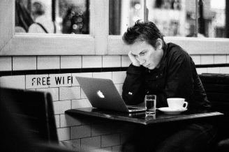 Free Wifi A person sits alone at a café table with a laptop, a cup of coffee, and a glass of water. The wall behind them has a "Free WiFi" sign. The person appears focused and leans their head on one hand.