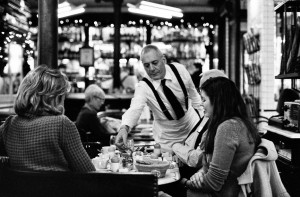 A waiter in a white shirt and black suspenders serves food to three women enjoying breakfast in Paris at a cozy, busy restaurant adorned with shelves of bottles and string lights. The lively scene is captured in classic black and white.