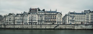 A row of historic, multi-story buildings with balconies lines the waterfront on L’île Saint Louis under an overcast sky in Paris, France. Leafless trees stand on the sidewalk in front of the stone embankment along the river at petit matin.
