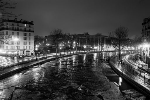 A black-and-white nighttime view of a partially frozen canal Saint Martin in Paris, with streetlights illuminating the scene. Buildings line both sides of the canal, and bare trees are visible. Reflections from the lights shimmer on the icy water surface as it gèle.