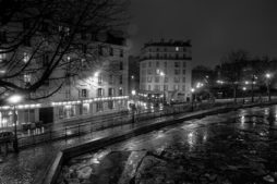 A black-and-white image of a quiet city street at night near Canal Saint Martin. Buildings are illuminated by streetlights, and bare trees line the sidewalk. Wet pavement and a reflective surface indicate recent rain. The scene exudes a serene, calm atmosphere.