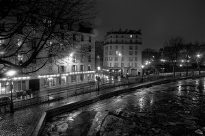 A black-and-white image of a quiet city street at night near Canal Saint Martin. Buildings are illuminated by streetlights, and bare trees line the sidewalk. Wet pavement and a reflective surface indicate recent rain. The scene exudes a serene, calm atmosphere.