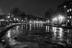 A black and white photograph of a nighttime urban scene with streetlights illuminating the icy surface of the canal Saint Martin. Bare trees line the canal, and buildings stand along both sides. Light reflections create a serene and moody atmosphere, capturing the essence of hiver as water gèle.