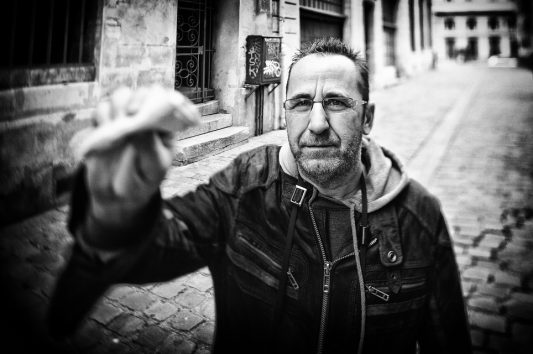 Lens Cleaning A man in glasses and a jacket stands on a cobblestone street, holding a lens cleaning cloth close to the camera. The image is black and white, with a blurred background of old buildings.
