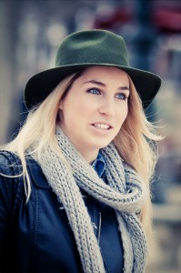 A woman with long blonde hair is wearing Le chapeau vert, a grey knitted scarf, and a black leather jacket. She is looking slightly to the right and appears to be outdoors in a city setting. The background is blurred with a mix of colors.