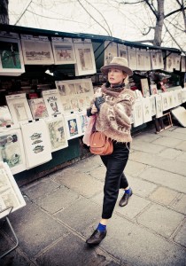A femme élégante in a hat, poncho, and gloves strolls past an outdoor art market displaying prints and paintings. She carries a shoulder bag among bare trees and stone pavement, embodying effortless style.
