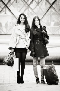 Two young women stand side by side outdoors, smiling at the camera. Après le Shopping, one wears a light coat, short skirt, and carries a handbag; the other sports a dark coat, jeans, and holds a large shopping bag.
