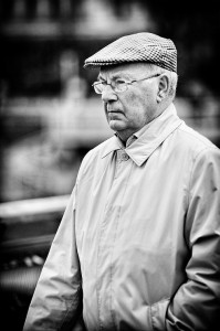 An elderly homme à la casquette, wearing glasses and a light-colored trench coat, stands outdoors looking thoughtful. The black and white photo features a blurred background, highlighting his contemplative mood.