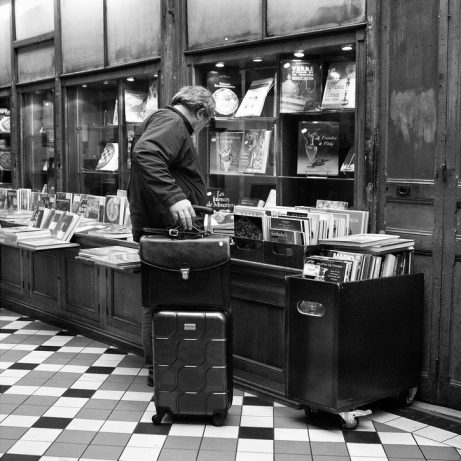 A man with a suitcase (homme à la valise) browses books inside a shop with checkered floor tiles and shelves filled with vieux livres and magazines. The scene is in black and white.