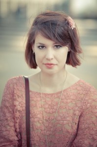 La jeune fille à la fleur, with short brown hair and a pink lace top, wears a delicate flower hair clip as she gazes directly at the camera with a neutral expression. The background is softly blurred.