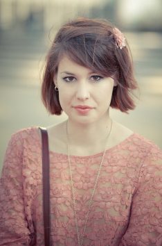 La jeune fille à la fleur La jeune fille à la fleur, with short brown hair and a pink lace top, wears a delicate flower hair clip as she gazes directly at the camera with a neutral expression. The background is softly blurred.