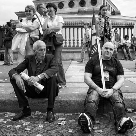 A black-and-white image of two men sitting on a stone curb in a public square during the manifestation on 23 septembre 2010. One man, dressed in a suit, holds a newspaper on his knee, while the other, in casual clothes and holding a flag, symbolizes the ongoing réforme des retraites. Several people stand in the background, some smiling and observing the scene.