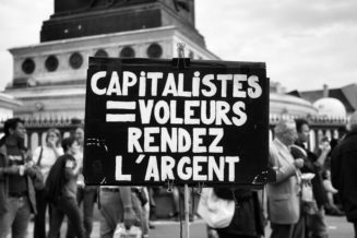 A protest sign in French reads "CAPITALISTES = VOLEURS, RENDEZ L'ARGENT" in white letters on a black background. The sign is held up during a manifestation on 23 septembre 2010, with people walking nearby and a monument visible in the background, highlighting the réforme des retraites issue.