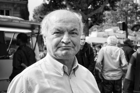 Black-and-white photo of an older man with a neutral expression, standing outdoors in a crowd. He is wearing a light-colored dress shirt. The image, taken during the 23 septembre 2010 manifestation against the réforme des retraites, shows trees, several people, a vehicle, and some building structures in the background.
