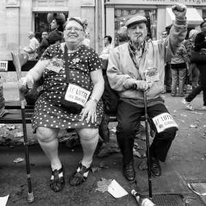 A black and white photo of an elderly woman and man sitting on a bench during the 23 septembre 2010 manifestation. The woman smiles, holding a water bottle and cane, while the man, wearing a jacket with a whistle in his mouth, raises a fist. Both have signs reading "Je lutte des classes" amid the réforme des retraites protests.