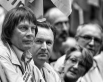 A black-and-white photo taken on 23 septembre 2010 shows four adults sitting closely together. The focus is on a man with medium-length hair on the left, wearing a collared shirt and jacket with a name tag. The other three adults, two men and one woman, are slightly out of focus in the background.