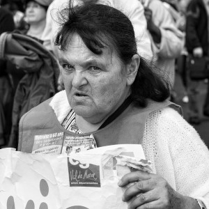 Black and white image of an elderly woman at a manifestation on 23 septembre 2010. She has a serious expression and is holding a sign. Wearing a light-colored cardigan and a vest with text about the réforme des retraites, she's surrounded by other people in the crowd.