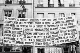 A large banner with French text is hung between buildings in a city street. The banner discusses topics like economic struggles, political critiques, and societal issues, addressing the impact of capitalism, unemployment, exclusion, and the erosion of democracy. It also references the Manifestation on 16 septembre 2010 against réforme des retraites.